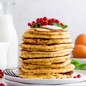 A stack of almond milk pancakes topped with red grapes, yogurt, and mint leaves, placed on two stacked white plates.