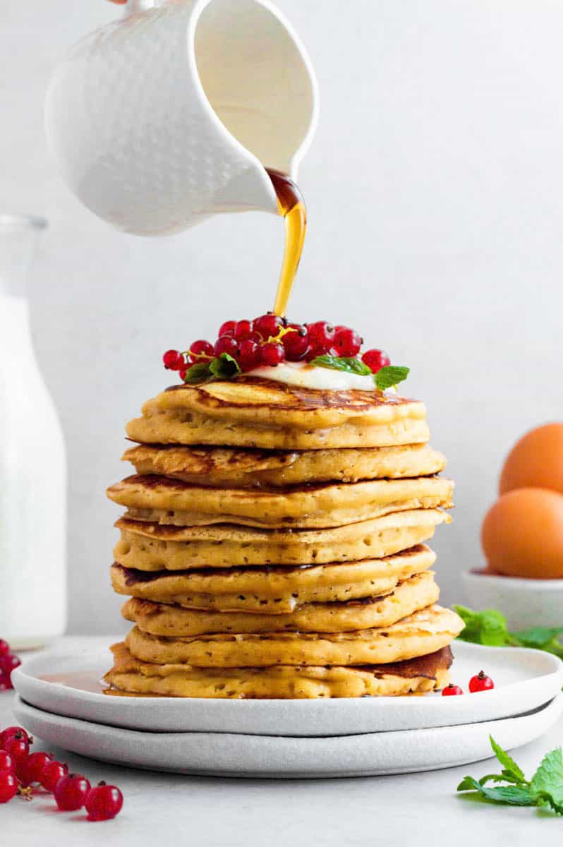 Pouring maple syrup from a small white pot over a stack of almond milk pancakes.