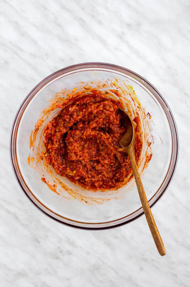 Shredded onion, minced garlic, ketchup, and spices mixed in a clear mixing bowl with a wooden spoon in it.