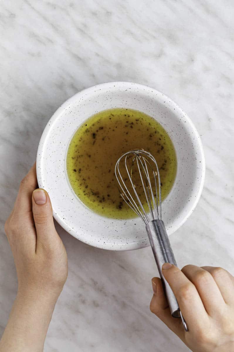Two hands holding a white bowl filled with salad dressing and mixing it with a stainless-steel whisk. 