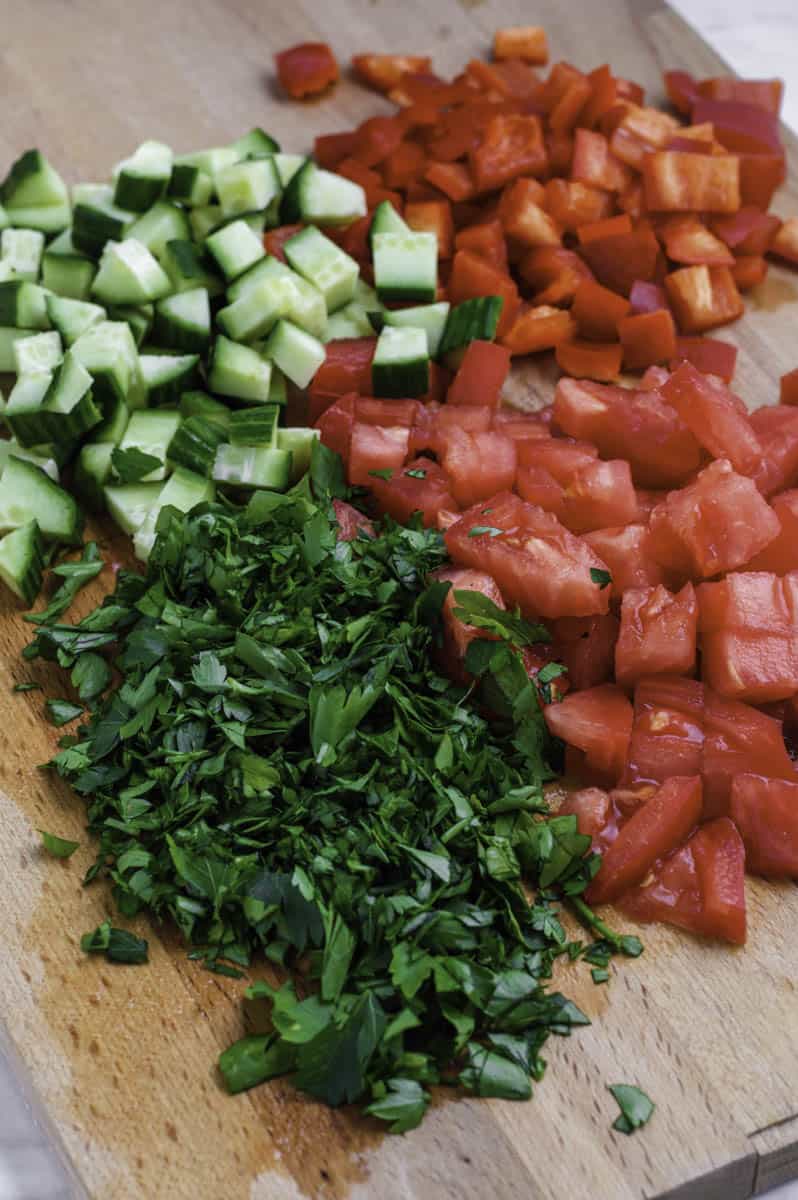 Chopped cucumber, tomato, red pepper, and parsley on a wooden cutting board.