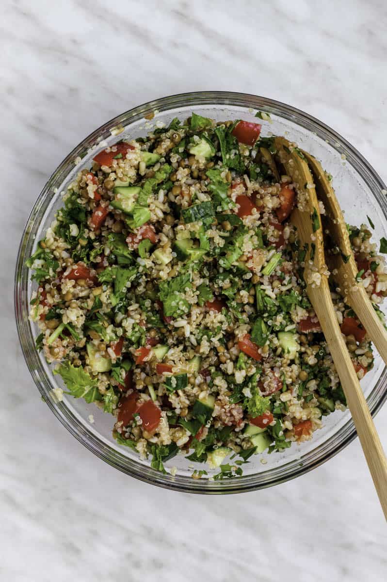 Mixed quinoa salad in a glass bowl with two wooden spoons in it.