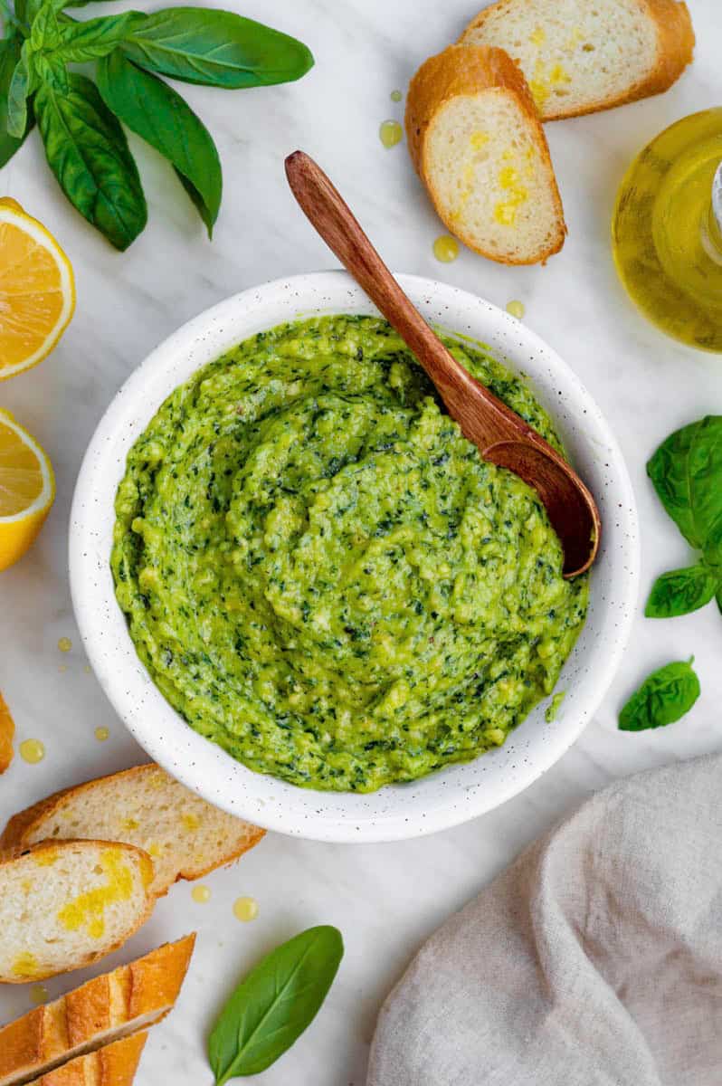 Cashew pesto served in a white bowl with a wooden spoon in it. Sliced baguette, basil leaves and halved lemon placed next to the bowl.