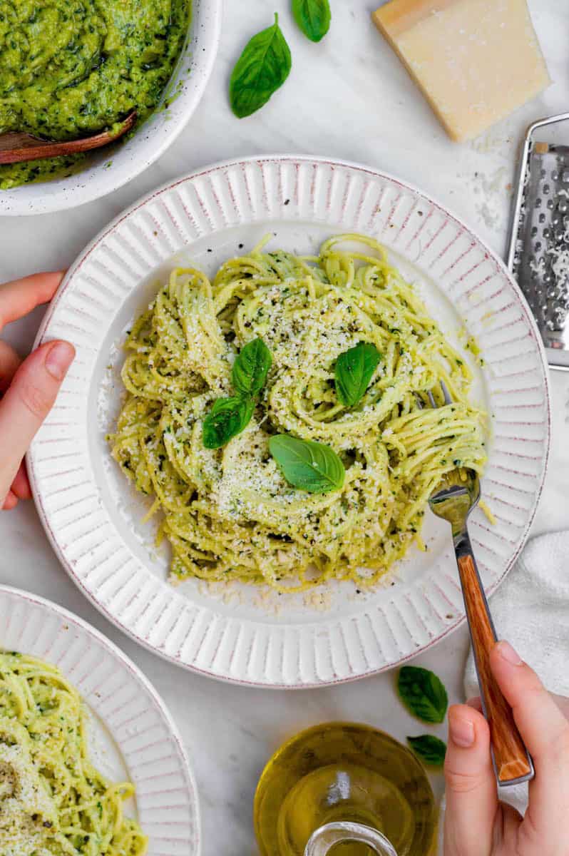 Pesto pasta served in a white plate and topped with grated parmesan and basil. A hand holding a fork that is inside the pasta.