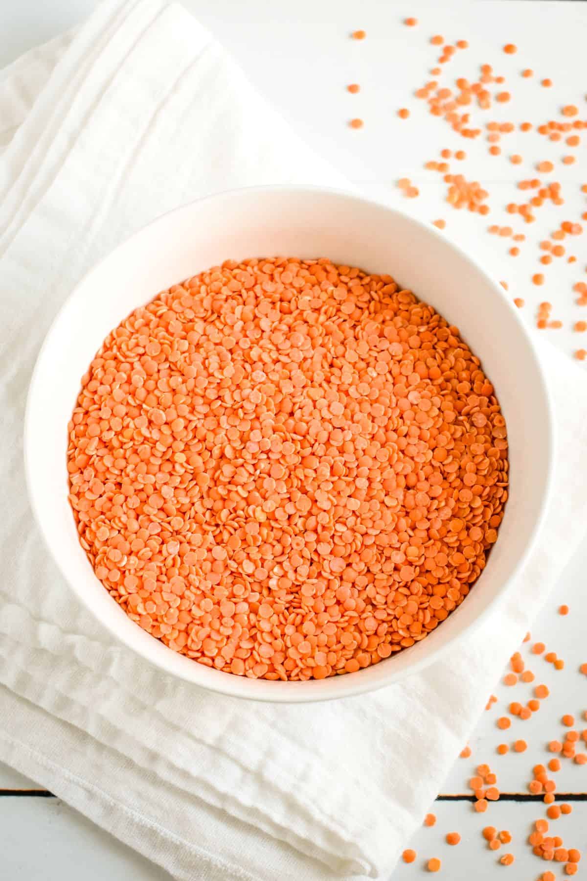 overhead of dry red lentils in a white bowl with some scattered around on the table.