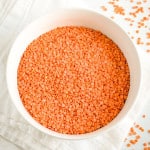 close-up of uncooked red lentils in a white bowl with some scattered around on the table.