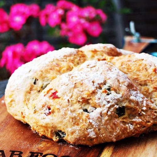 Homemade bread with bits of peppers and veggies baked into it on wood with flowers behind.