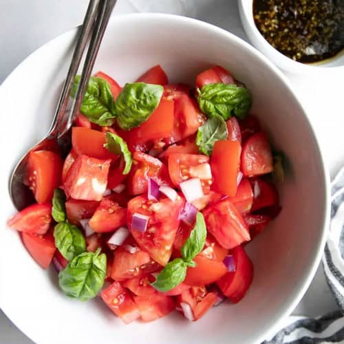 Bowl of tomatoes with bits of red onion and small basil leaves served with balsamic vinaigrette.