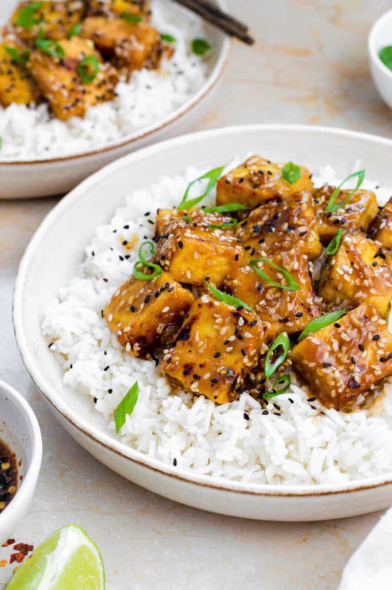 Honey garlic tofu served over cooked white rice and topped with black sesame seeds and chopped green onion.