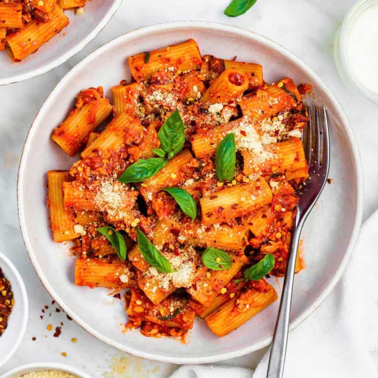 A white plate filled with tofu pasta topped with vegan parmesan and basil leaves. A silver fork placed on the right side of the plate.