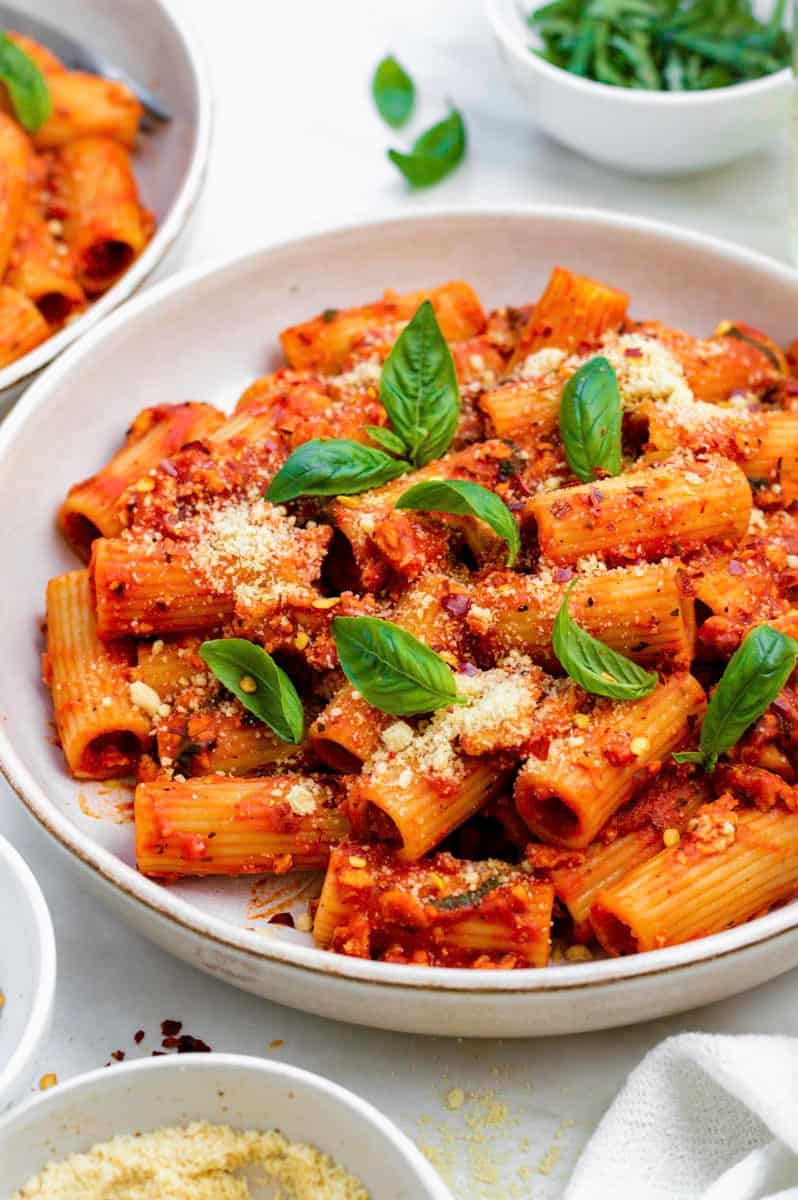 Pasta tomat tahu disajikan di piring putih dan di atasnya ditaburi basil, parmesan vegan, dan serpihan paprika merah.