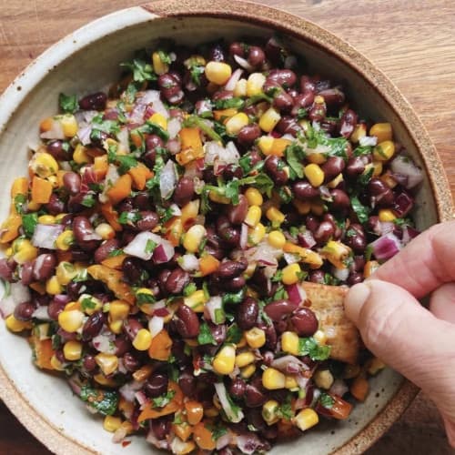 Black bean and vegetable salad in a wooden bowl with a hand dipping a cracker in it.