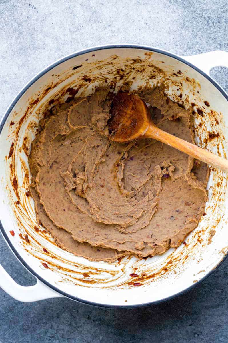 Refried beans in a white Dutch oven with a wooden spoon placed in it.