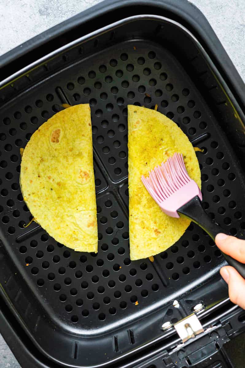 Two quesadillas in the air fryer basket. A hand brushing one of the quesadillas with oil using a pastry brush.