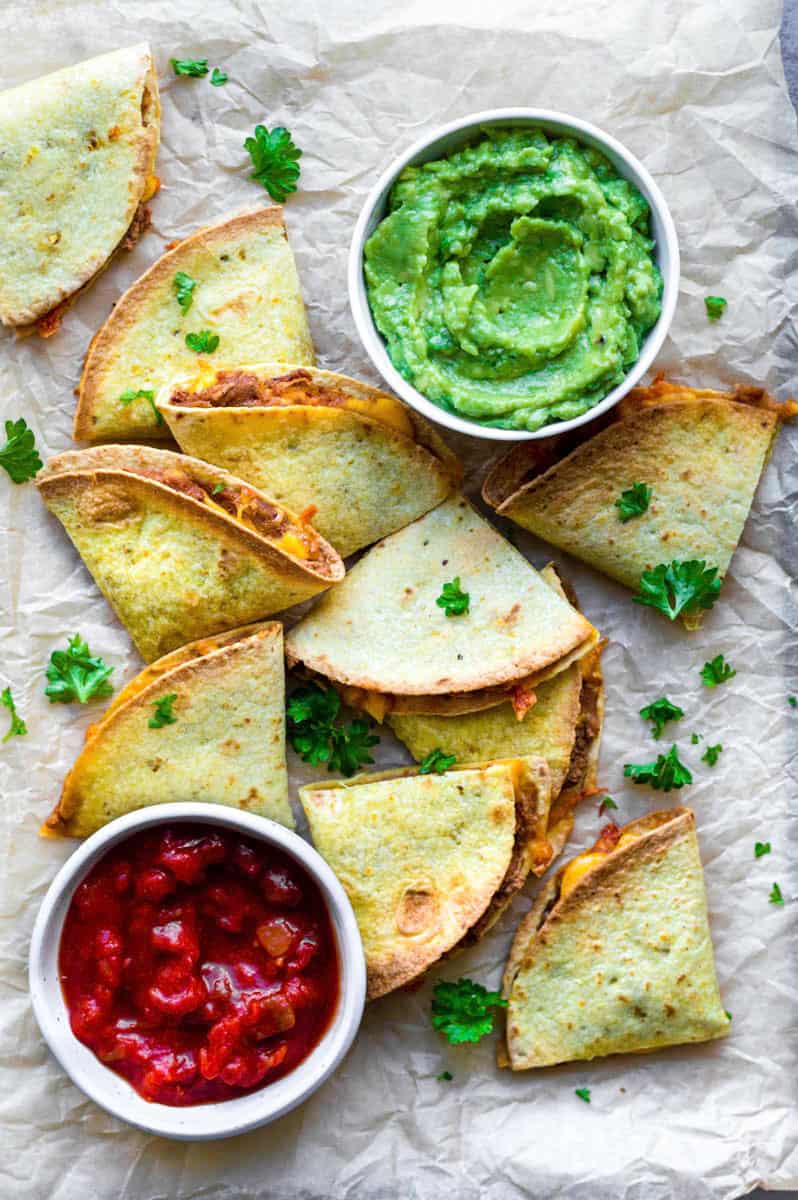Air fryer quesadillas served on a plate lined with parchment paper and topped with parsley.  A small bowl of guacamole is placed on the right side of the quesadillas, and a small bowl of salsa is placed on the left.