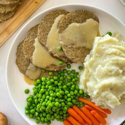 Vegan meat and gravy on a plate with mashed potatoes, peas and carrots.