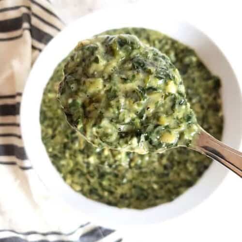 Creamy spinach in a spoon held up over white serving bowl.