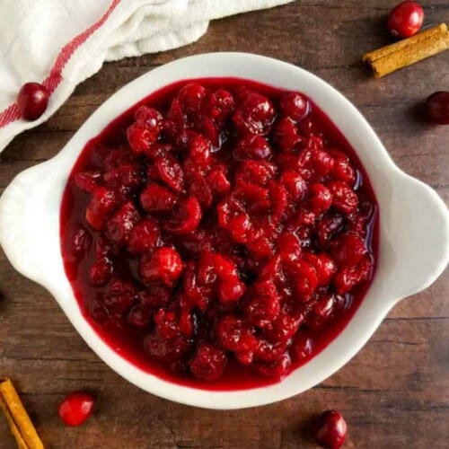 Bright red cranberry sauce in a bowl with fresh cranberries and cinnamon sticks on the table.