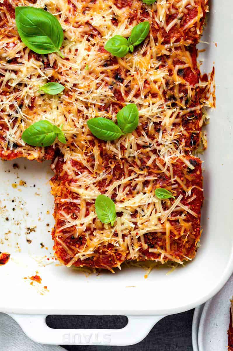 Vegan tofu parmesan in a white baking dish with a pieces cut off from it on the left, and topped with fresh basil.