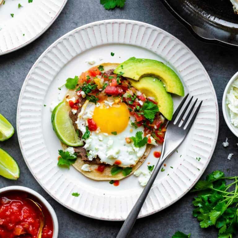 Vegetarian Huevos Rancheros served on a white plate and topped with feta cheese, avocado, and fresh cilantro.