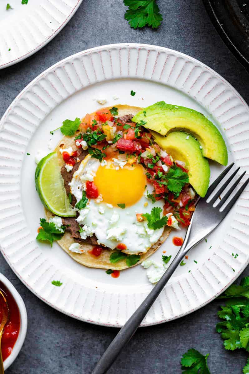 A white plate with vegetarian huevos rancheros served inside.  Topped with feta cheese, avocado and fresh coriander.  A silver fork on the right side of the plate.