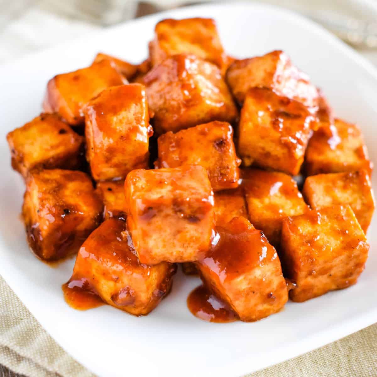close-up of bbq tofu pieces on a white plate with sauce dripping off of them.