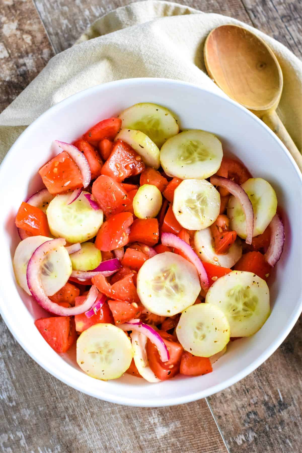 overhead of cucumber and tomato salad in a white bowl with a wooden spoon next to it on a kitchen napkin.