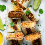Halved burritos served on a white board layered with parchment paper, topped with fresh cilantro. Lime wedges placed on both sides of the burritos.