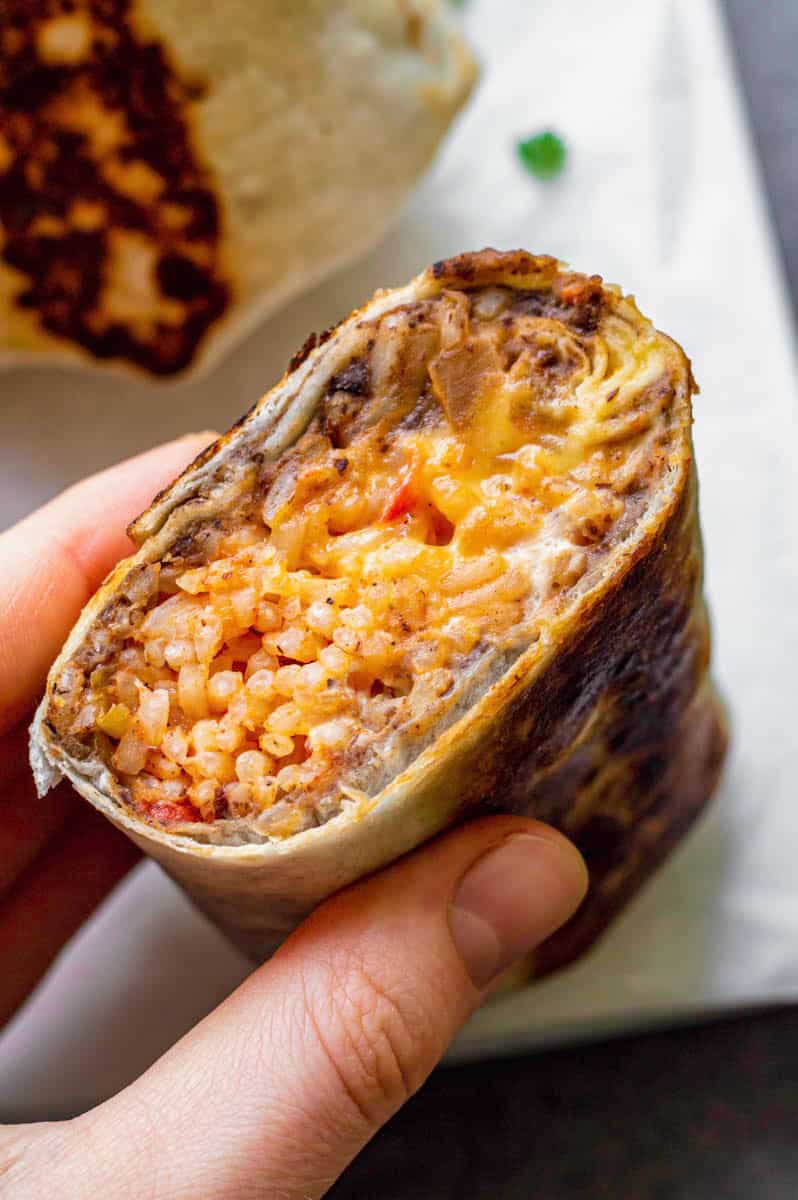 A hand holding half a burrito filled with rice, beans and cheese sauce.
