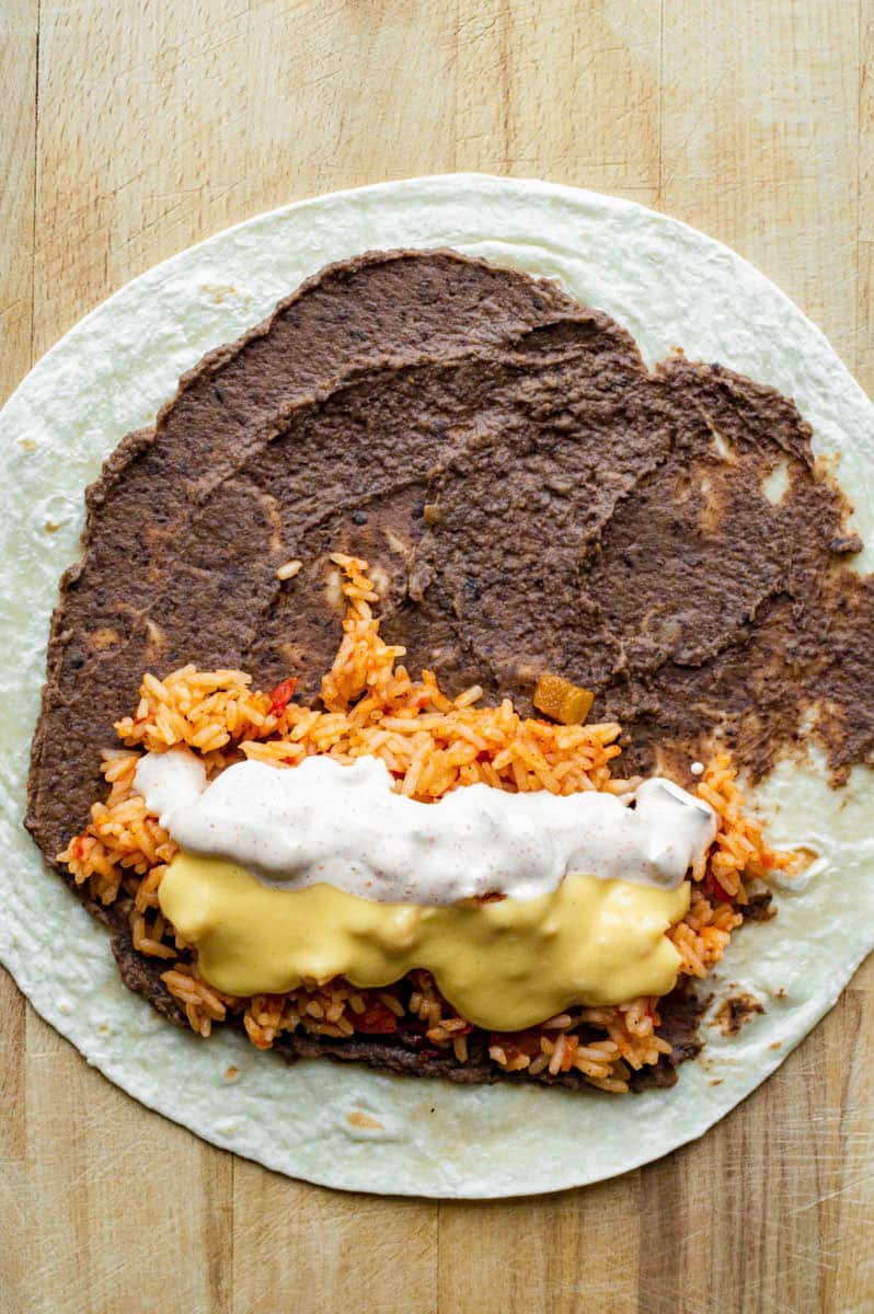 A flour tortilla placed on a wooden cutting board and top with refried beans, rice, cheese sauce, and jalapeno sauce.