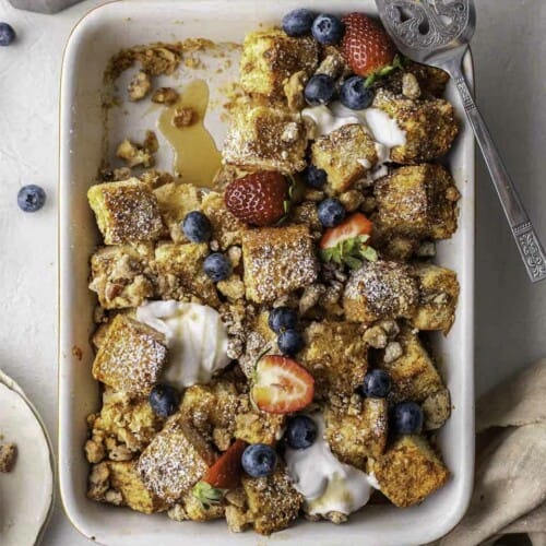 Vegan French toast cubes baked in a casserole dish. The dish is dusted with powdered sugar and is topped with a pecan crumble, berries and cream.