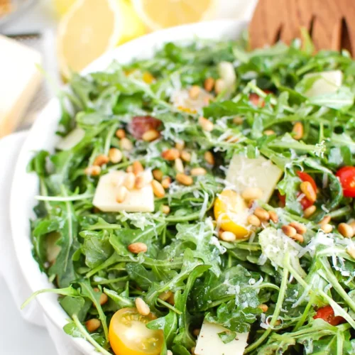 lemon arugula salad in a white bowl with a wooden salad claw in it.