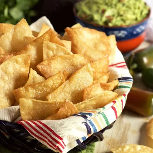 tortilla chips in a basket with a kitchen towel in it and some guacamole in the background.
