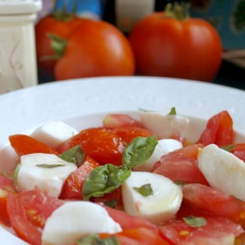 close-up of caprese salad in a white bowl witih tomatoes behind it.