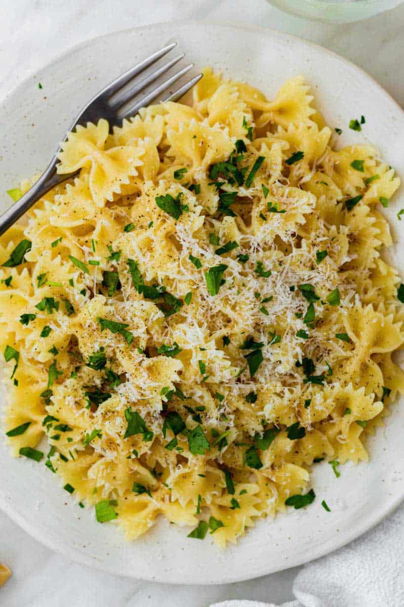 A close-up photo of cooked farfalle pasta served in a white plate and topped with parmesan cheese and parsley.