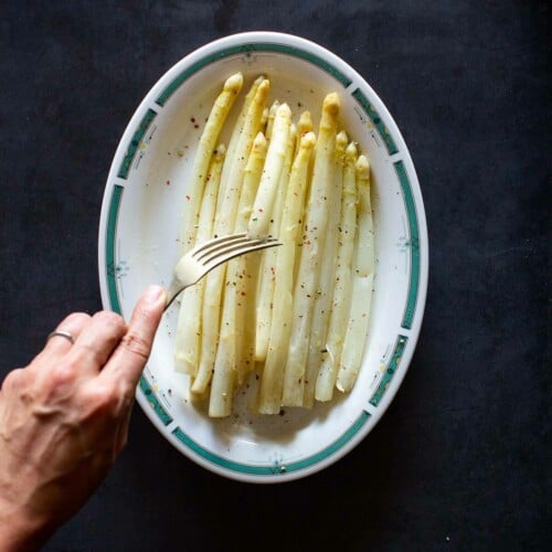overhead of white asparagus in a dish with hand sticking fork in one.