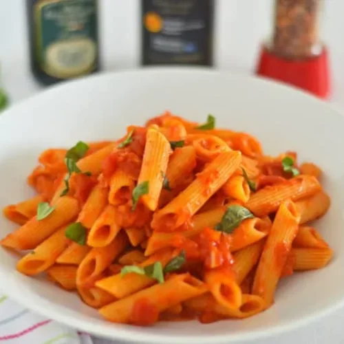 arrabiata pasta in a white bowl with a fork on a kitchen napkin next to it.