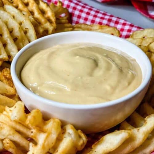 White bowl full of creamy light orange sauce in a bed of waffle fries.