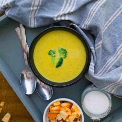 Vegan Broccoli Cheddar Soup in a blue bowl with croutons, spoons and a towel around it.