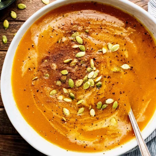 Orange autumn squash soup in a white bowl with pumpkin seeds and cinnamon on top, with a spoon in the bowl.