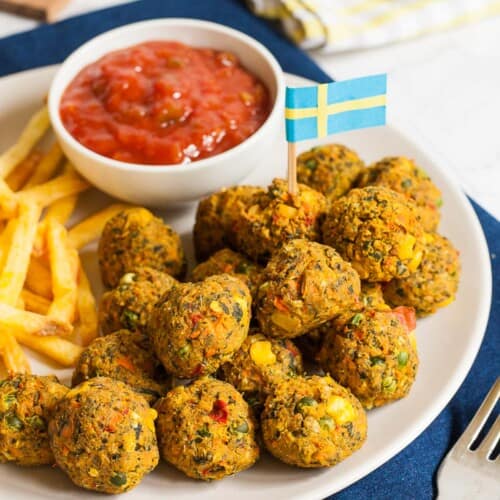 White plate full of small colourful balls with fried potato sticks and a small white bowl with red sauce. A blue yellow flag is stuck in one ball.