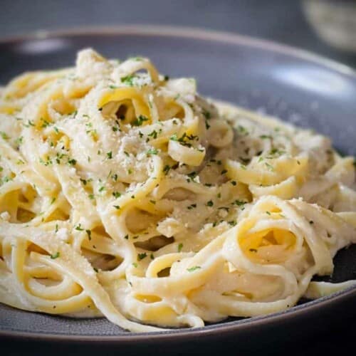 A pile of fettuccini Alfredo on a grey plate, topped with parmesan cheese and chopped parsley.