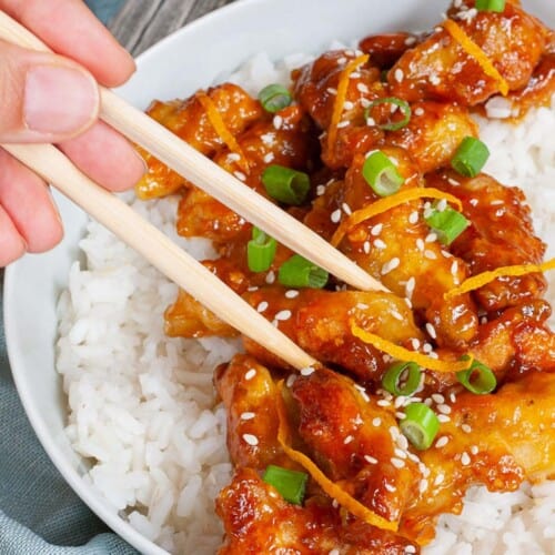 A large white bowl with white rice and sticky brown battered strips on top sprinkled with sesame seeds, orange peels and chopped spring onion. A hand is holding chopsticks and taking on strip.