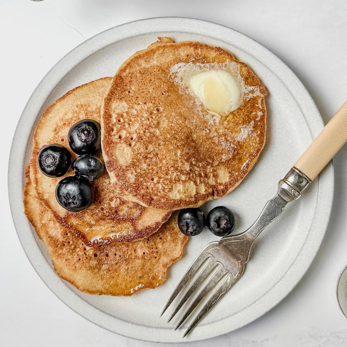 Top dairy-free pancakes with sliced ​​butter and blueberries and serve on a plate with a fork next to it.