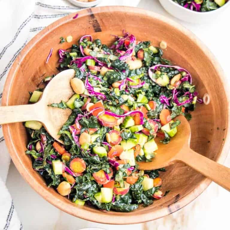 A large wooden bowl filled with chopped Tuscan kale salad and balsamic miso dressing, two wooden spoons next to it.