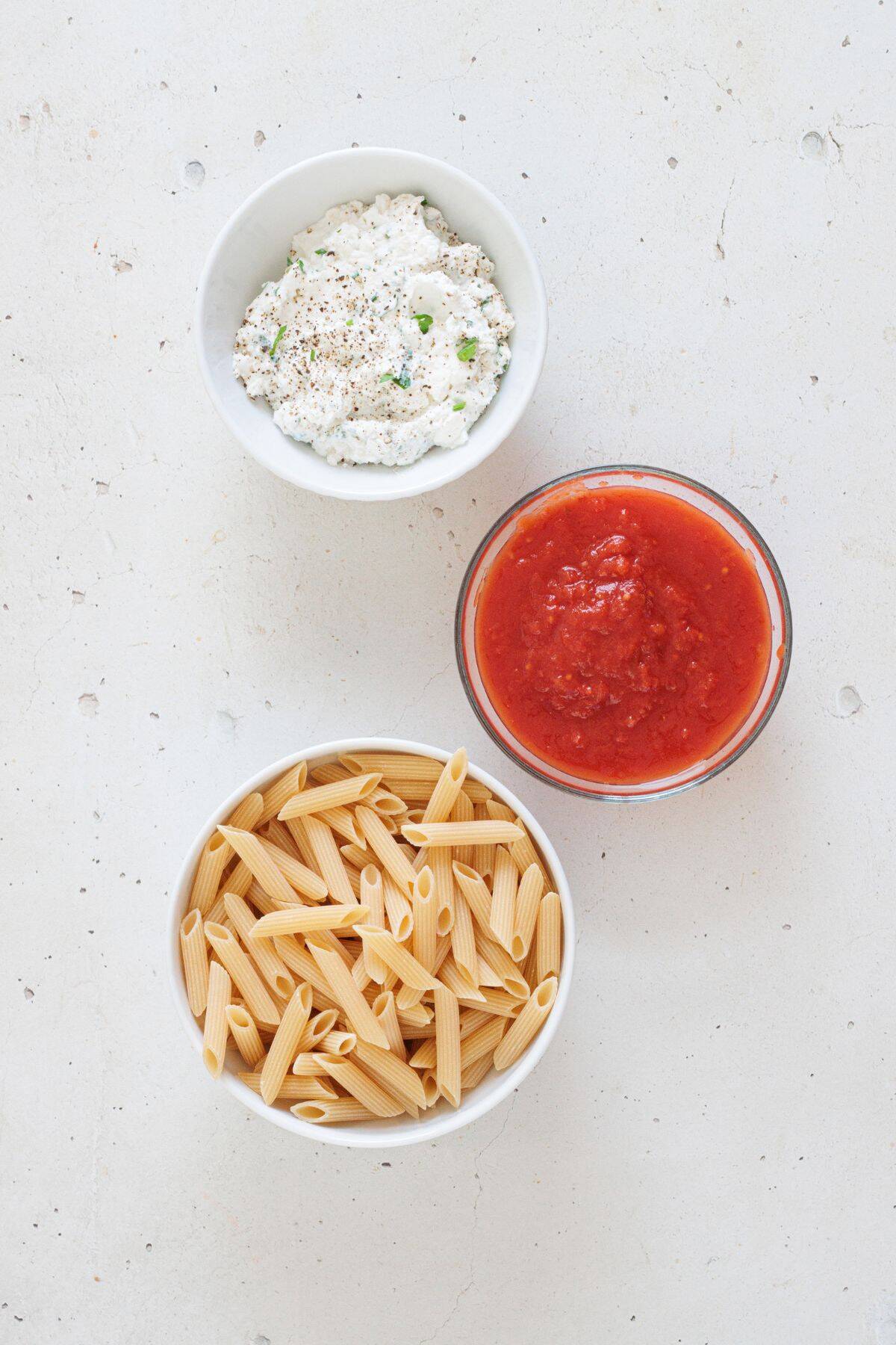 Ingredients to make vegan baked ziti including sunflower seed ricotta, canned tomato sauce, and penne pasta
