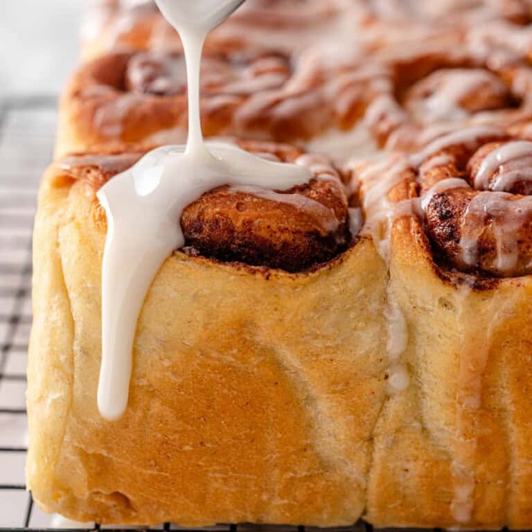 A spoon drizzling vanilla icing onto the corner of the warm vegan cinnamon rolls batch on a wire cooling rack