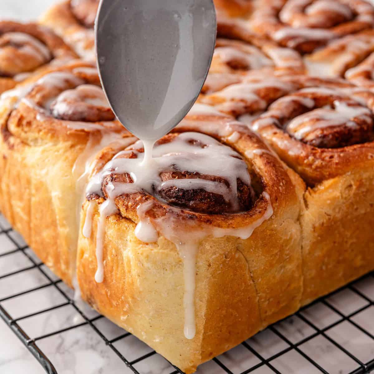 A spoon drizzling vanilla glaze onto freshly baked vegan cinnamon rolls on a wire cooling rack, showing the first application of icing
