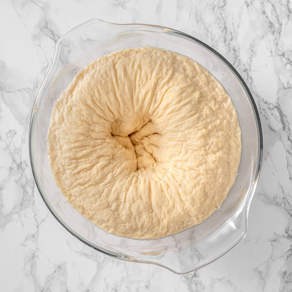 Vegan cinnamon roll dough punched down after rising, showing the deflated, wrinkled surface in the glass bowl