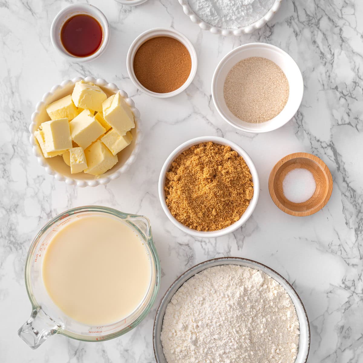 Overhead flat lay of all vegan cinnamon roll recipe ingredients measured in bowls on a marble surface: flour, brown sugar, vegan butter, plant milk, cinnamon, yeast, vanilla, and salt